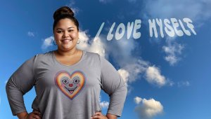 A confident person stands beneath a blue sky with scattered clouds, wearing a shirt with a colorful heart and smiling face. Above them, the words “I LOVE MYSELF” appear in cloud-like letters—symbolizing self-love and emotional wellness.