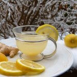 Glass cup of lemon ginger tea with citrus slices and fresh ginger on a white plate, set against a snowy outdoor background.