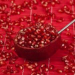 Halved pomegranate with spoon and scattered seeds on a red textured surface, highlighting the fruit’s vibrant color and nutrient-rich appeal.