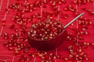Halved pomegranate with spoon and scattered seeds on a red textured surface, highlighting the fruit’s vibrant color and nutrient-rich appeal.