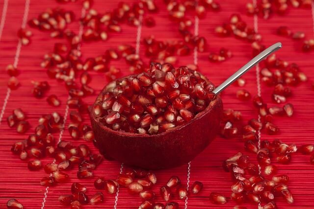 Halved pomegranate with spoon and scattered seeds on a red textured surface, highlighting the fruit’s vibrant color and nutrient-rich appeal.