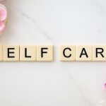 Scrabble-style tiles spelling “SELF CARE” on a white marble surface, surrounded by soft pink and white flowers.