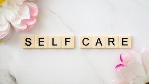 Scrabble-style tiles spelling “SELF CARE” on a white marble surface, surrounded by soft pink and white flowers.