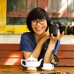 Person sitting outdoors at a rustic wooden table, smiling while holding a Nikon camera, with a teapot and teacup in front.