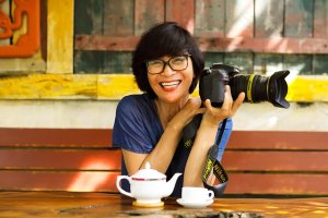 Person sitting outdoors at a rustic wooden table, smiling while holding a Nikon camera, with a teapot and teacup in front.